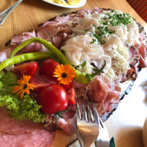 Ein Fest für die Sinne! Auf einer rustikalen Holzplatte sind köstliche Jausenplatten kunstvoll arrangiert. Verschiedene Sorten von Käse, Wurst, frischem Brot, knackigem Gemüse und süßen Früchten laden zum Schlemmen ein. Die Vielfalt der Aromen und Texturen verspricht eine wahre Geschmacksexplosion.